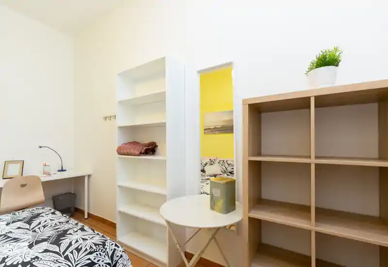 Bright, neatly arranged bedroom corner with patterned bedding, shelving and a small desk — ideal for a single occupant. The room feels fresh with a simple, modern setup and good natural light.