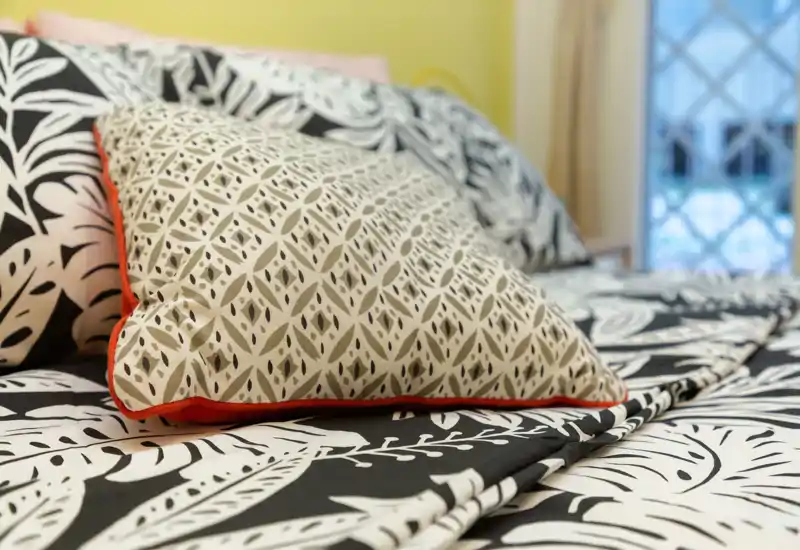 Primo piano di un letto ben rifinito con biancheria stampata e cuscini decorativi, luce naturale luminosa dalla finestra e atmosfera accogliente e moderna.