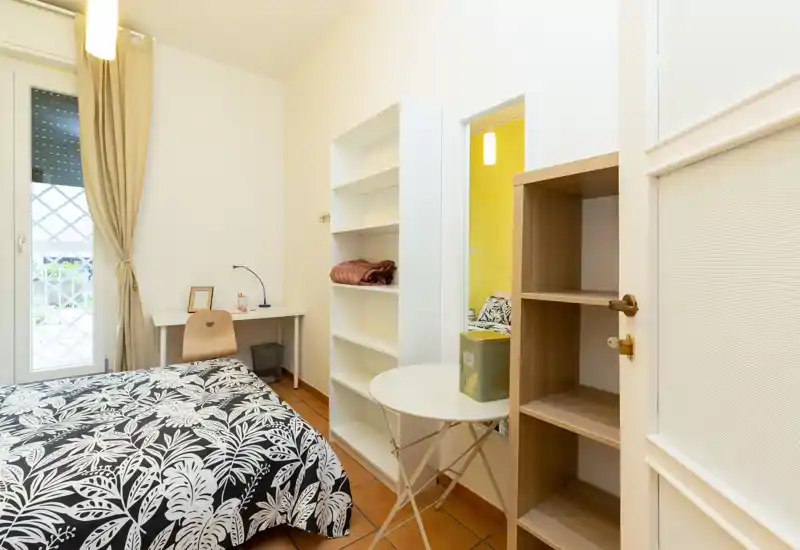 Bright, tidy bedroom with a patterned double bed, small desk by the window and open shelving — ideal for a single tenant or couple.
