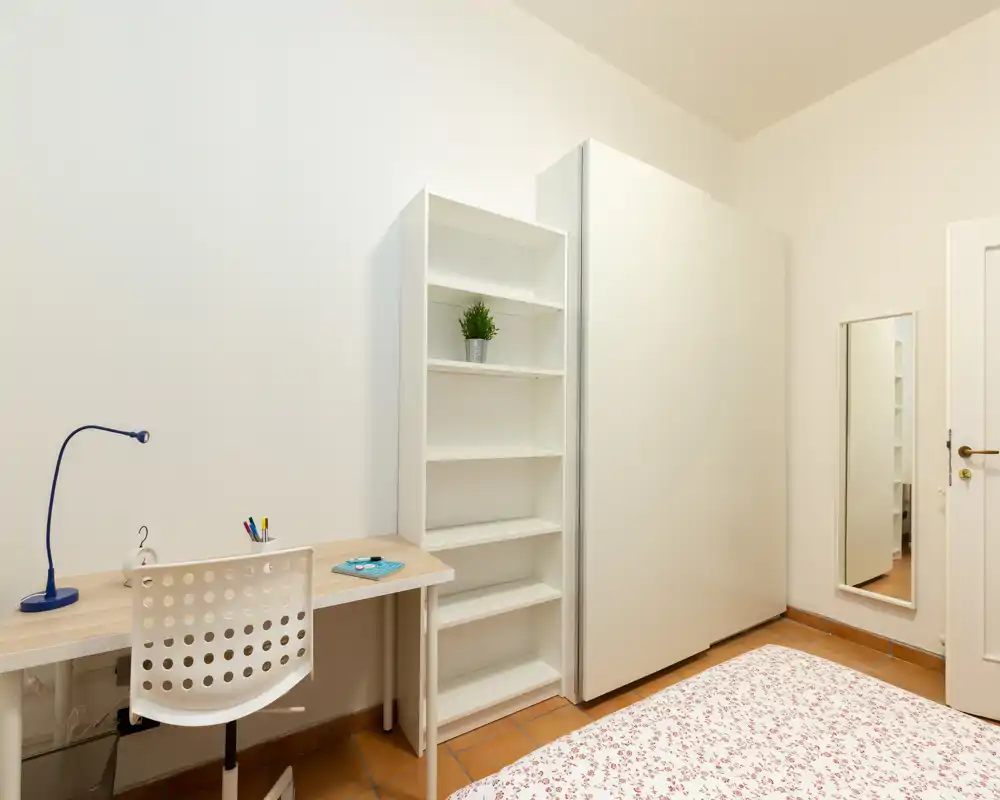 Minimal, well-lit bedroom corner with a partially visible bed, a desk and shelving — ideal for a student or single occupant seeking a clean, functional space.