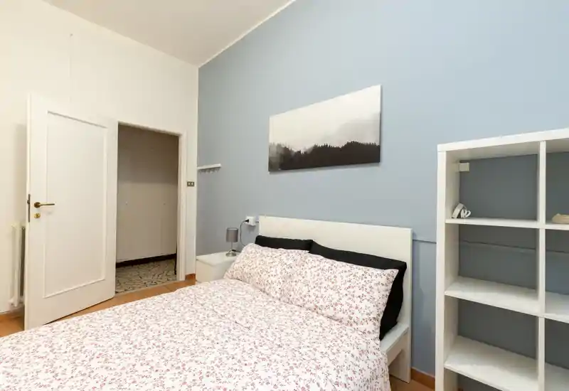 Dormitorio luminoso y bien presentado con cama doble con ropa de cama estampada, mesita con lámpara, cuadro y estantería abierta — perfecto para anuncios de alquiler que destacan confort y decoración moderna.