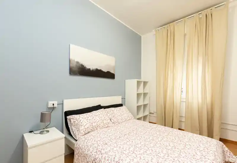 Dormitorio luminoso y ordenado con cama doble con ropa de cama estampada, mesita con lámpara y estantería sencilla. Tonos neutros y luz natural suave crean un ambiente tranquilo y acogedor, perfecto para un anuncio de alquiler.