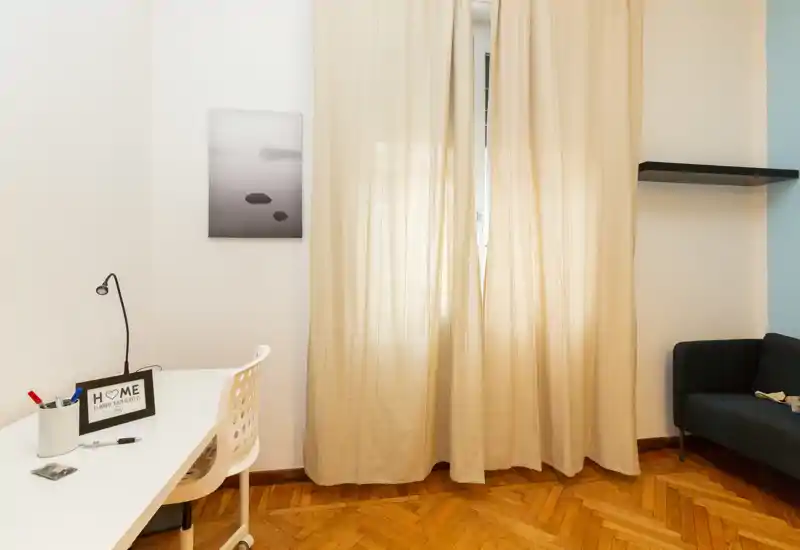 Bright, simply styled living area with a small desk, chair and a compact sofa beside large curtains — ideal for remote work and relaxing.