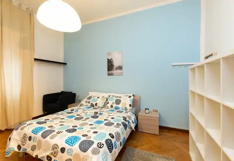 Dormitorio luminoso y bien decorado con cama doble, funda nórdica estampada, mesita y estanterías; pared azul y suelo de parquet que aportan calidez.