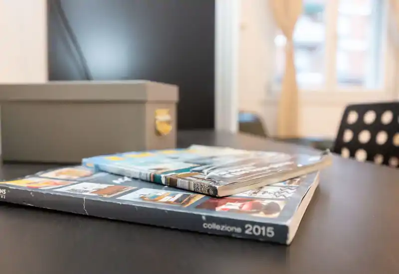 Close-up of stacked magazines/books on a dark table surface; background hints at an interior (chair, window, curtains) but the image focuses on the reading materials rather than a specific room.