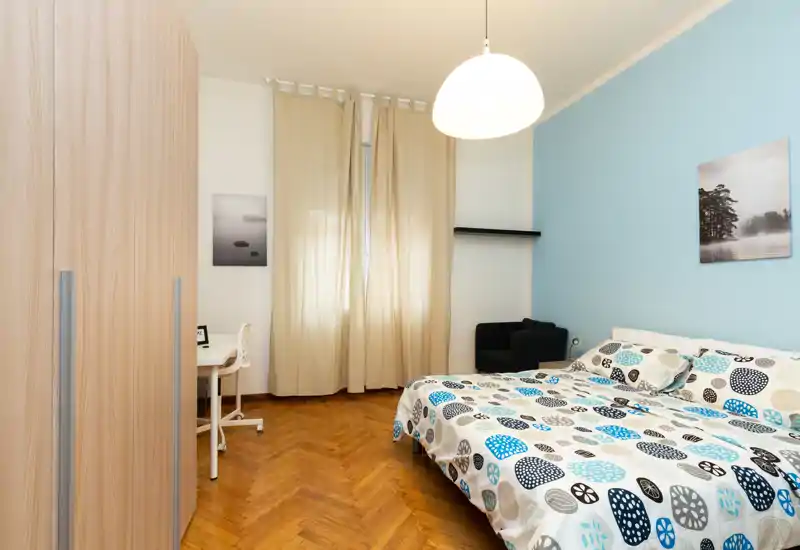 Camera da letto luminosa e ben arredata con letto matrimoniale coperto da una trapunta fantasia, armadio, scrivania e poltrona su una parete azzurro tenue.