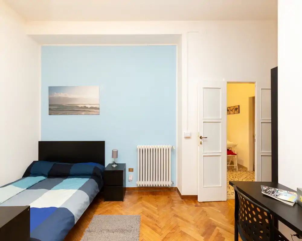 Bright, neatly staged bedroom with a double bed against a soft blue accent wall, bedside table and lamp, parquet floor and a small desk near the doorway.