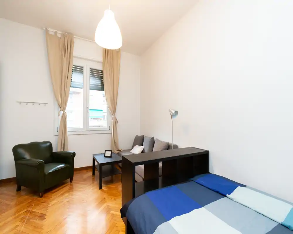 Bright, simply furnished bedroom with a double bed in the foreground, a small sofa and coffee table, and a comfortable armchair near the window.