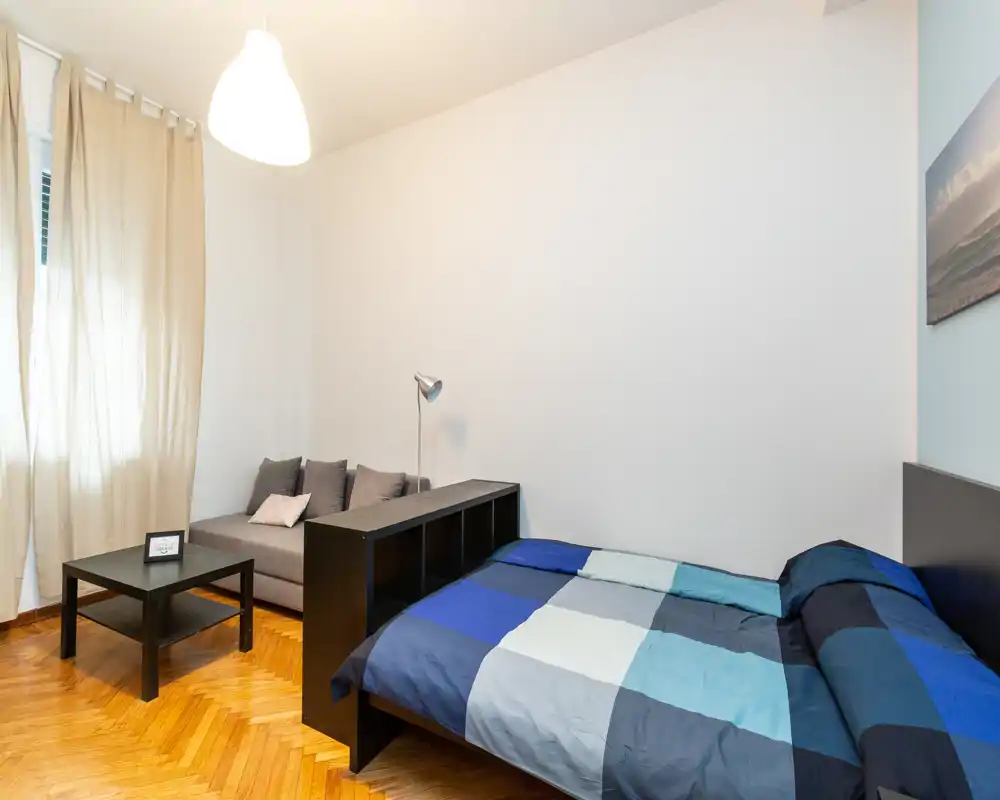 Bright, modern bedroom with a double bed covered in blue geometric bedding, a small sofa and coffee table, and warm parquet flooring.