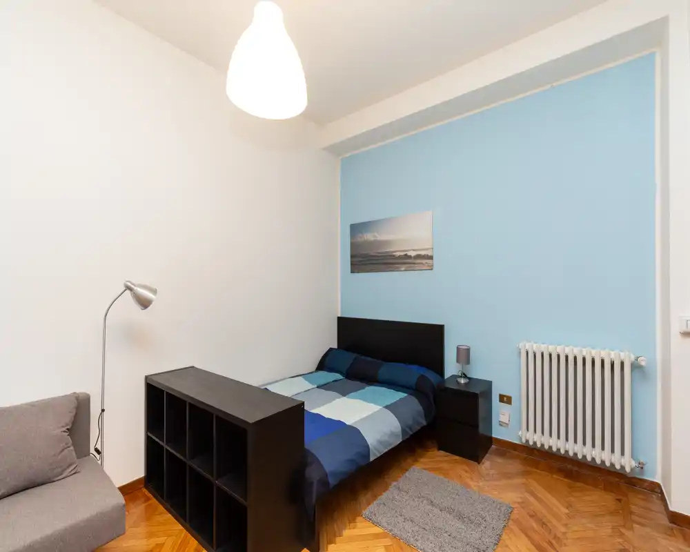 Comfortable, well-lit bedroom with a double bed against a soft blue accent wall, bedside table and lamp, decorative picture and hardwood floors — ideal for a cozy stay.