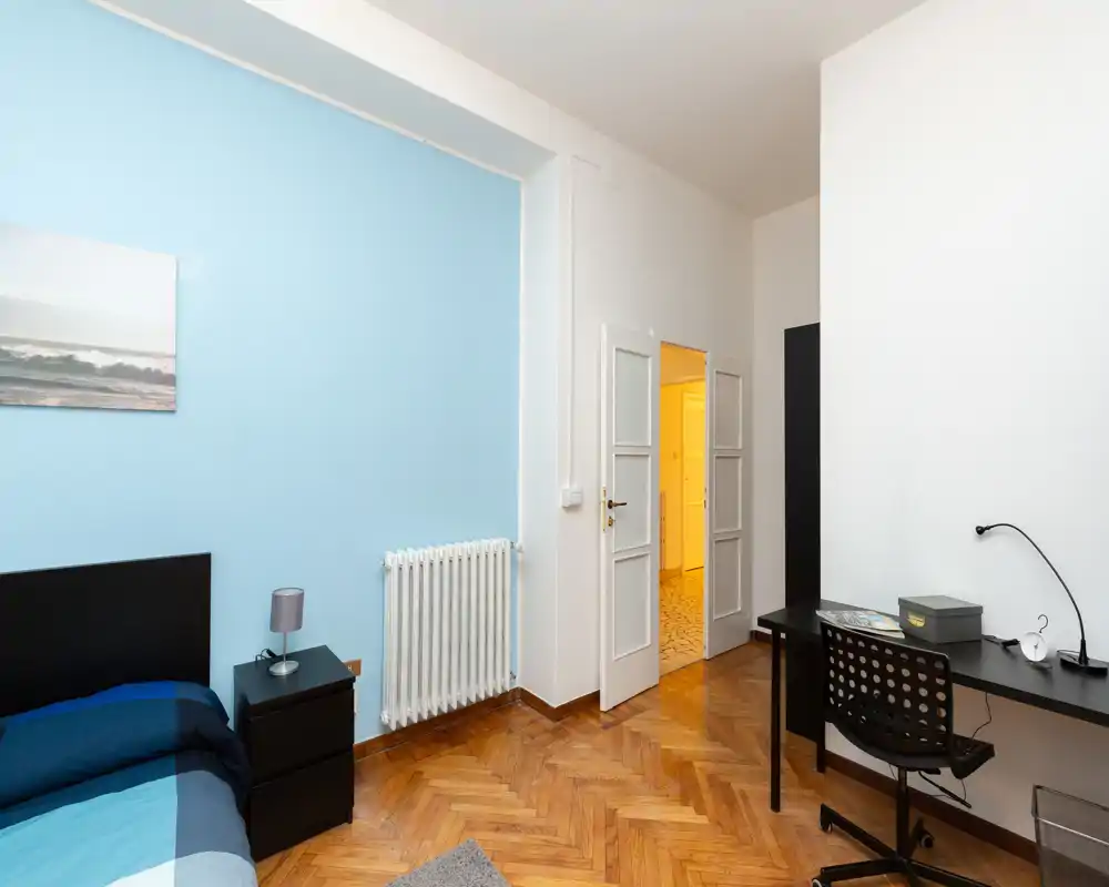 Neat, modern bedroom with a single bed against a pale blue accent wall, bedside table with lamp, radiator and a desk workspace; warm parquet flooring and soft, even lighting.