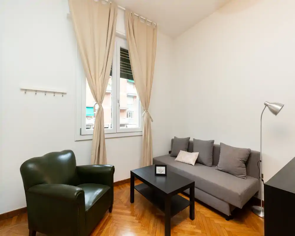 Bright, simply furnished living area with a grey sofa, accent armchair, coffee table and natural light from a window — ideal for relaxing in a compact apartment.