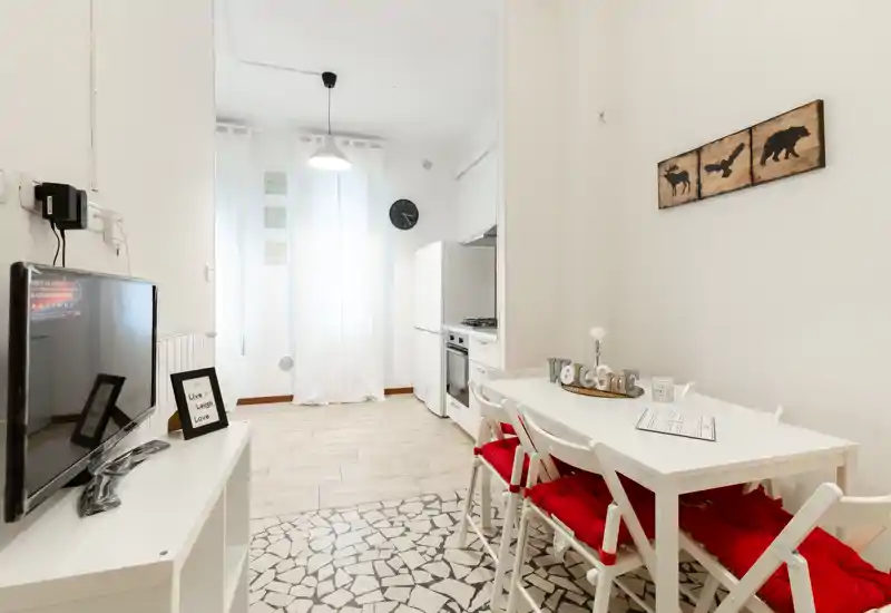 Zona giorno luminosa e accogliente con tavolo da pranzo e angolo cottura compatto. Pareti bianche, cuscini rossi e dettagli decorativi donano un aspetto fresco e invitante, perfetto per soggiorni brevi.