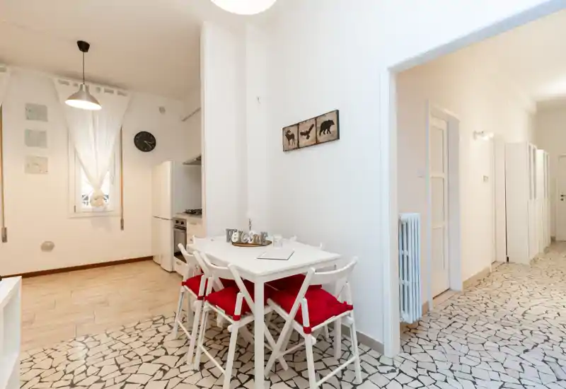Zona pranzo luminosa e ordinata con tavolo bianco e quattro sedie con cuscini rossi; cucina compatta visibile sullo sfondo e arredo murale di gusto.