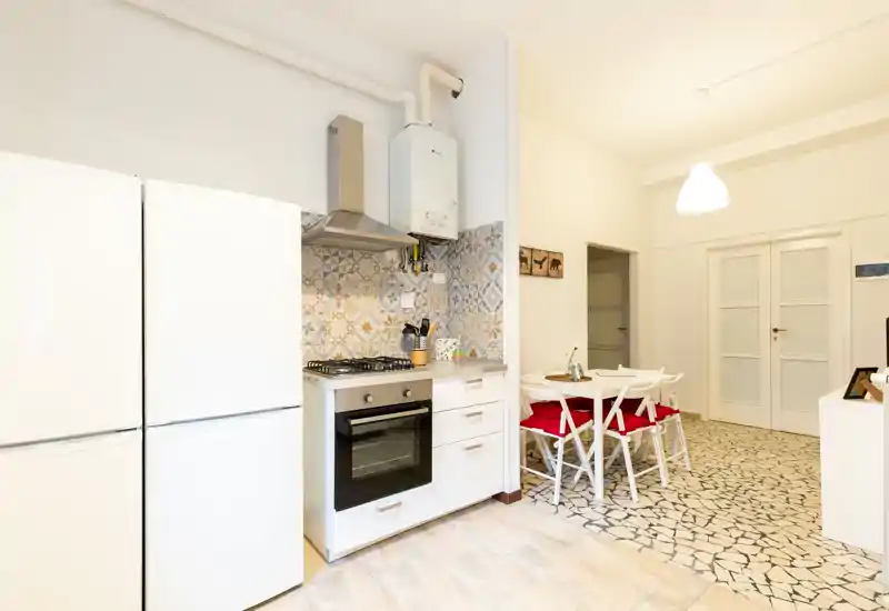 Cucina moderna e luminosa con rivestimento a piastrelle decorato, piano cottura a gas e forno incorporato; piccolo tavolo da pranzo con sedie dai cuscini rossi per un angolo colazione accogliente.