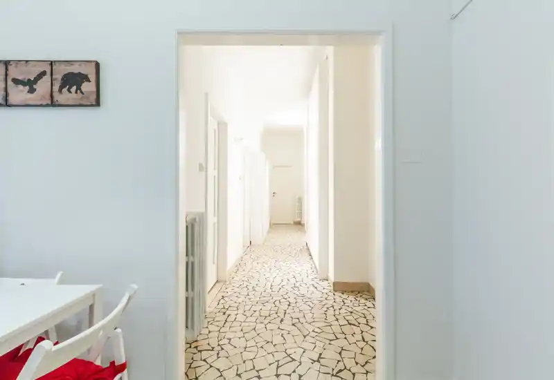 Ingresso/corridoio luminoso e pulito con pavimento effetto mosaico e pareti bianche; visuale su un corridoio lungo che collega le stanze.