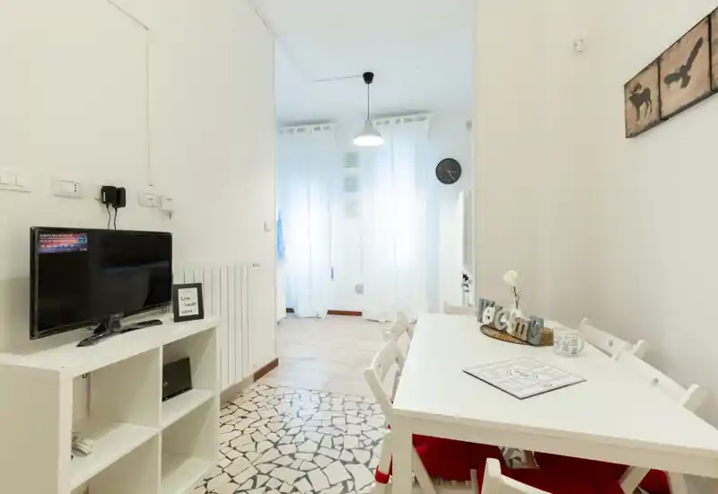 Soggiorno/pranzo luminoso e moderno con tavolo e sedie bianche, TV su mobile basso e luce naturale soffusa dalle tende trasparenti — accogliente e ben arredato per un annuncio.