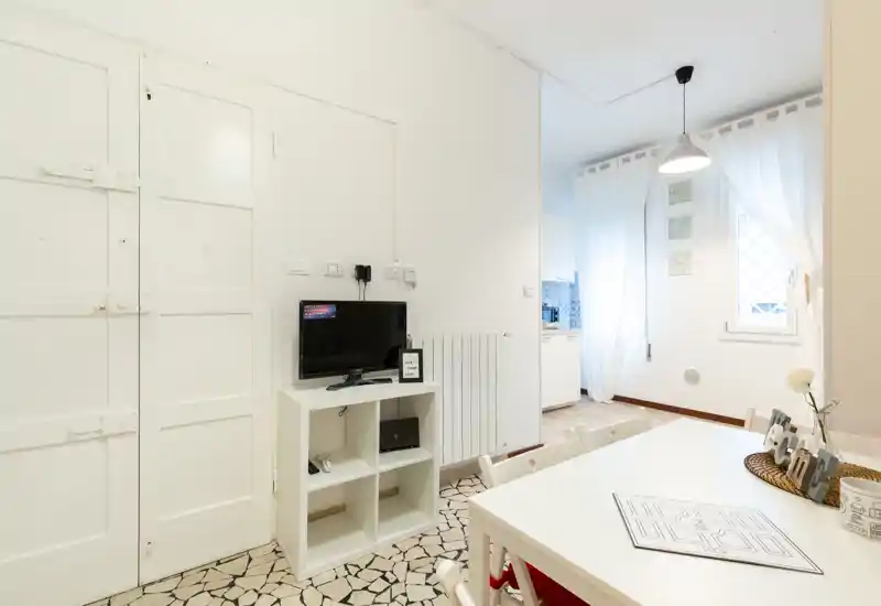 Zona giorno luminosa e minimale con piccolo mobile TV, tavolo da pranzo bianco e sedie, illuminazione naturale morbida dalla finestra con tenda.