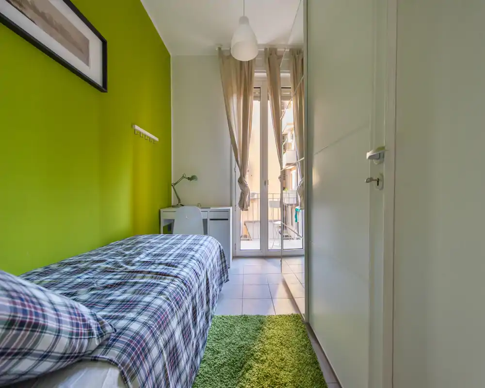 Dormitorio individual luminoso con cama de cuadros, pared verde y pequeño escritorio junto a la puerta del balcón — bien iluminado y ordenado para una estancia cómoda.