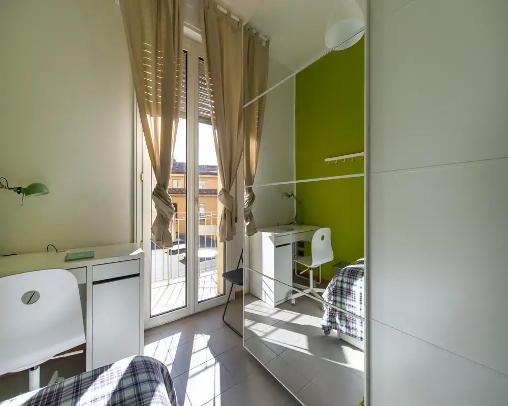 Dormitorio luminoso y compacto con cama individual, escritorio y amplio armario con espejo que multiplica la luz. Puertas balconeras altas ofrecen abundante luz natural y un cómodo rincón de estudio.