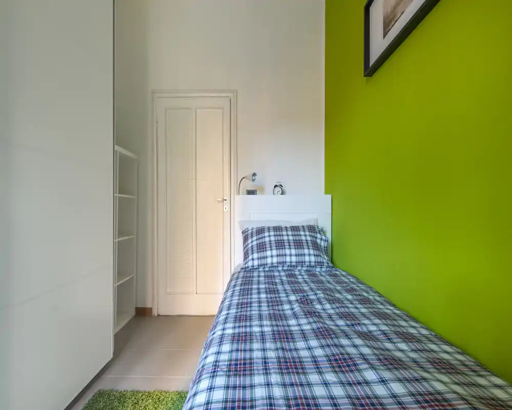 Dormitorio individual compacto y luminoso con cama de estampado a cuadros junto a una pared verde intensa, cabecero blanco, armario y lámpara de mesita — ideal para estudiante o alquiler corto.