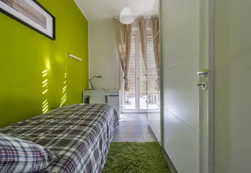 Dormitorio individual luminoso con pared verde, ropa de cama de cuadros, un pequeño escritorio junto a las puertas del balcón y armario empotrado. La luz natural atraviesa las cortinas ligeras, creando un espacio fresco y acogedor ideal para estudiantes o estancias cortas.