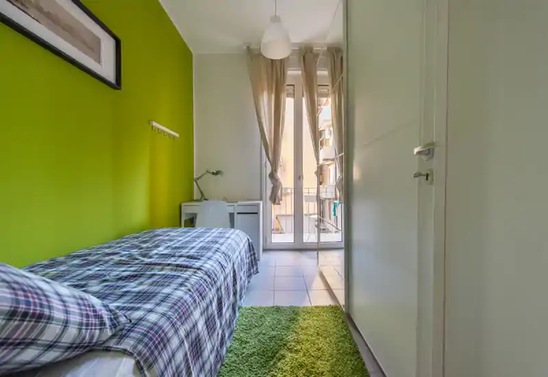 Dormitorio individual luminoso con cama de cuadros, pared verde y pequeño escritorio junto a la puerta del balcón — bien iluminado y ordenado para una estancia cómoda.