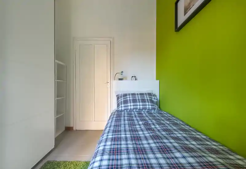 Dormitorio individual compacto y luminoso con cama de estampado a cuadros junto a una pared verde intensa, cabecero blanco, armario y lámpara de mesita — ideal para estudiante o alquiler corto.
