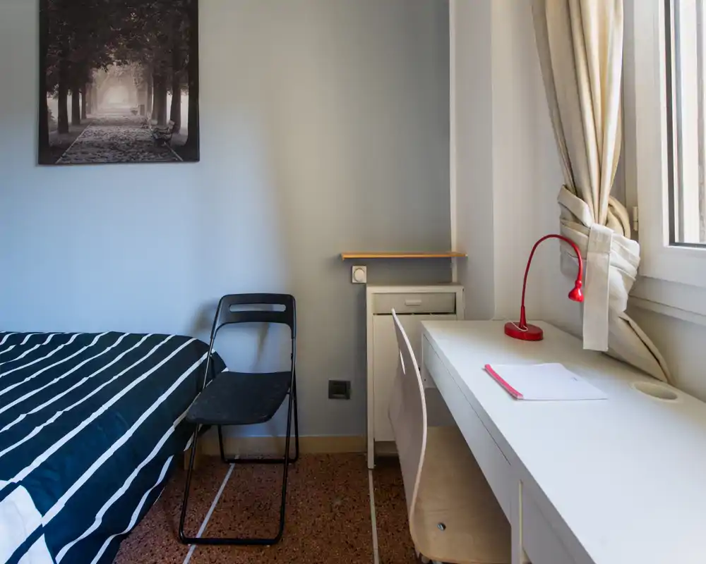 Dormitorio luminoso y sencillo con cama de rayas, escritorio junto a la ventana y lámpara de lectura roja — ideal para una persona o estudiante.