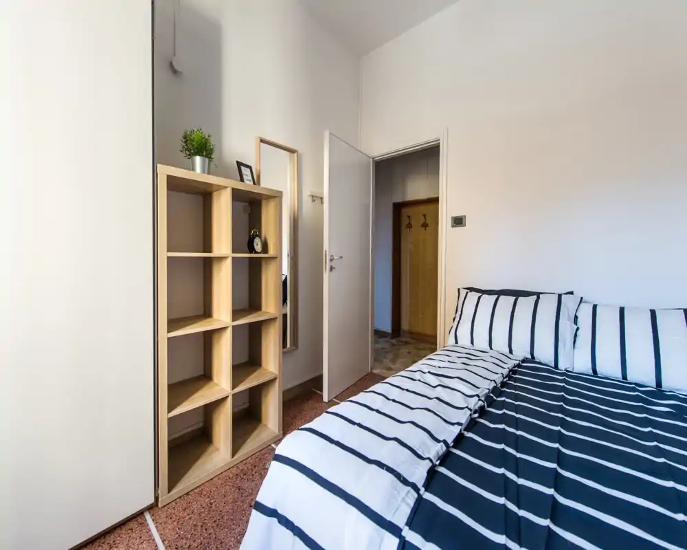Dormitorio luminoso y minimalista con cama doble de rayas y estantería abierta; paredes neutras y luz natural para un espacio limpio y acogedor, perfecto para alquileres.