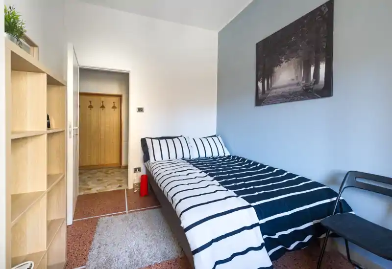 Dormitorio luminoso y ordenado con cama doble con ropa de cama a rayas, cuadro sobre la cabecera, pequeña alfombra y estantería para almacenamiento — ideal para un alquiler cómodo y compacto.