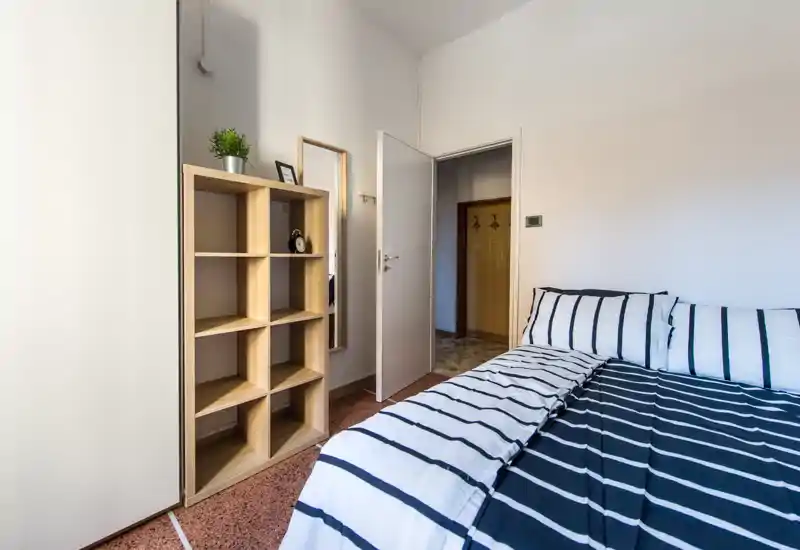 Bright, simply styled bedroom featuring a striped double bed and open shelving; neutral walls and natural light create a clean, welcoming space ideal for short-term rentals.