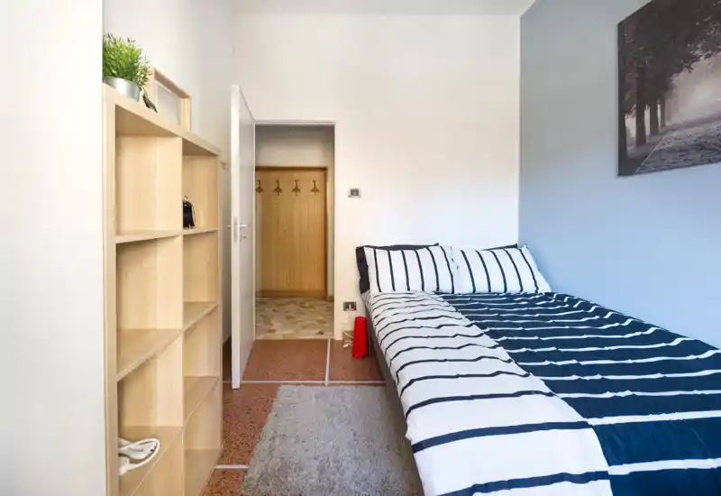 Dormitorio individual luminoso y ordenado con ropa de cama a rayas, estantería y alfombra; diseño moderno y luz natural, ideal para anuncios de alquiler.