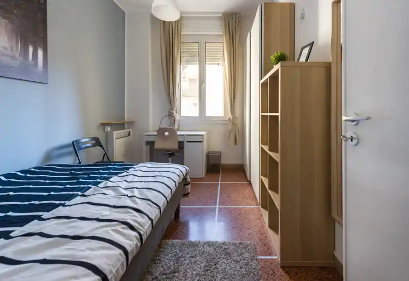 Dormitorio individual luminoso con cama cómoda, escritorio junto a la ventana y estanterías de madera; tonos neutros y una alfombra mullida crean un espacio tranquilo y ordenado, ideal para un estudiante o una sola persona.