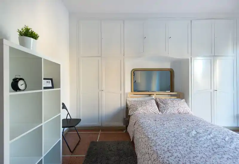 Dormitorio luminoso y acogedor con cama doble flanqueada por armarios empotrados blancos y un espejo decorativo; la ropa de cama estampada y la alfombra añaden calidez.