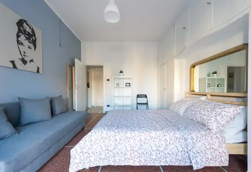 Dormitorio luminoso y ordenado con cama doble cubierta con edredón estampado, un gran espejo sobre la cabecera y un sofá gris junto a la pared — acogedor y bien iluminado.