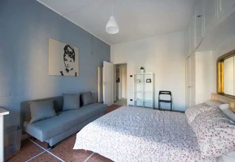 Camera da letto luminosa e di stile con letto matrimoniale con biancheria a motivo, divano grigio, armadi a muro e luce naturale morbida—ideale per un soggiorno confortevole.