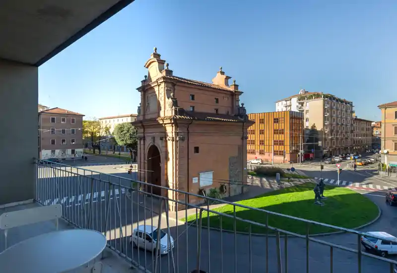 Sunny balcony with metal railing and small outdoor table, overlooking a historic city gate and a roundabout — ideal for morning coffee with an urban view.
