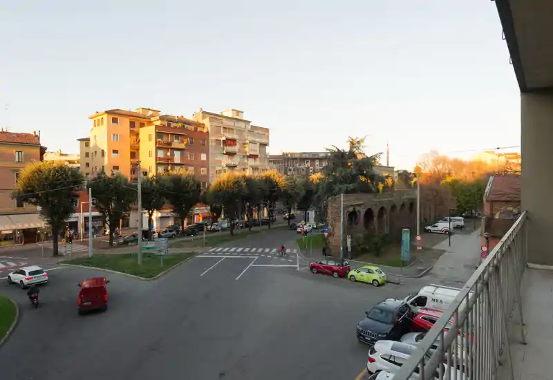 Balcony view overlooking a city intersection with parked cars, trees and low-rise apartment buildings bathed in warm late-afternoon light.