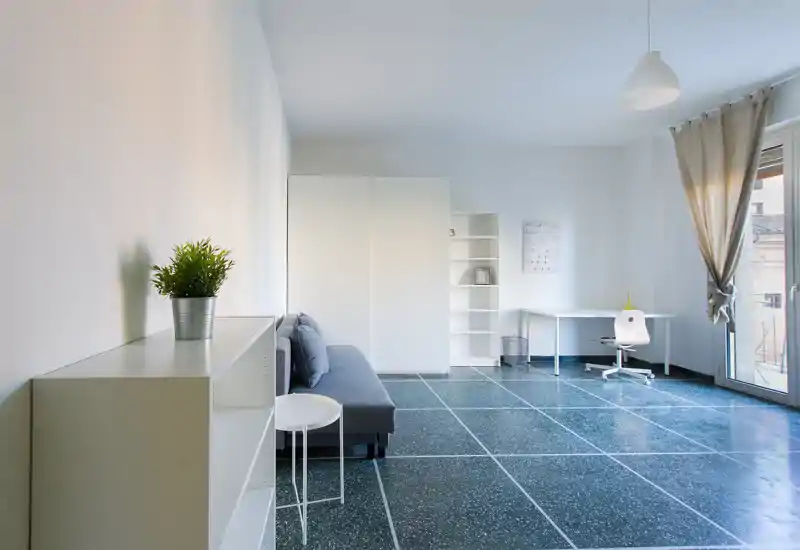 Zona giorno luminosa e minimal con divano compatto, scaffalatura e scrivania vicino a una grande finestra — perfetta per rilassarsi e lavorare in uno spazio moderno e arioso.