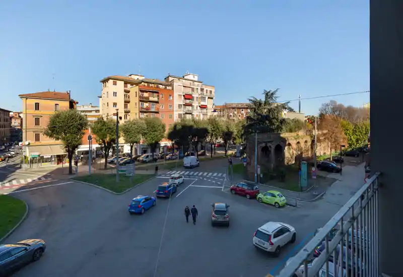 Wide daytime street view from a balcony showing a roundabout, parked cars, pedestrians and surrounding buildings under clear blue sky.
