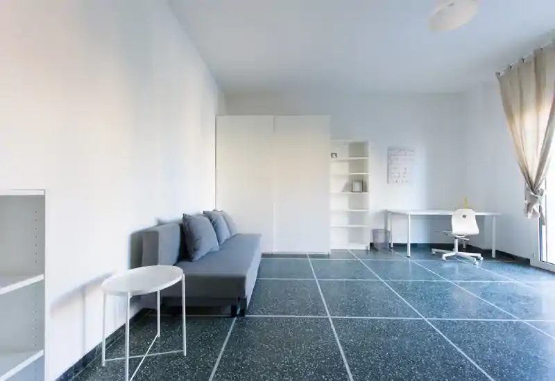 Bright, minimalist living area with a compact grey sofa, small side table, shelving and a workspace by a large window — ideal for a modern studio layout.
