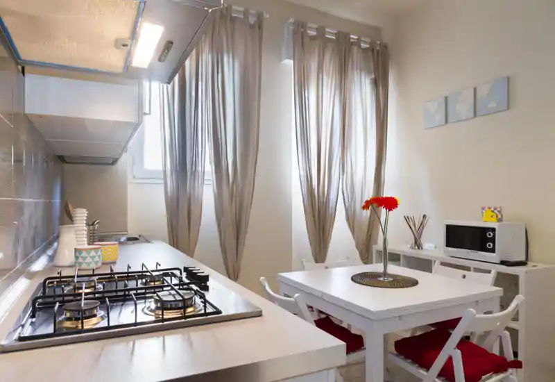 Bright, modern kitchenette with a gas hob integrated into the counter, a small dining table with red cushions and a fresh flower, and compact appliances—ideal for a cozy studio apartment.
