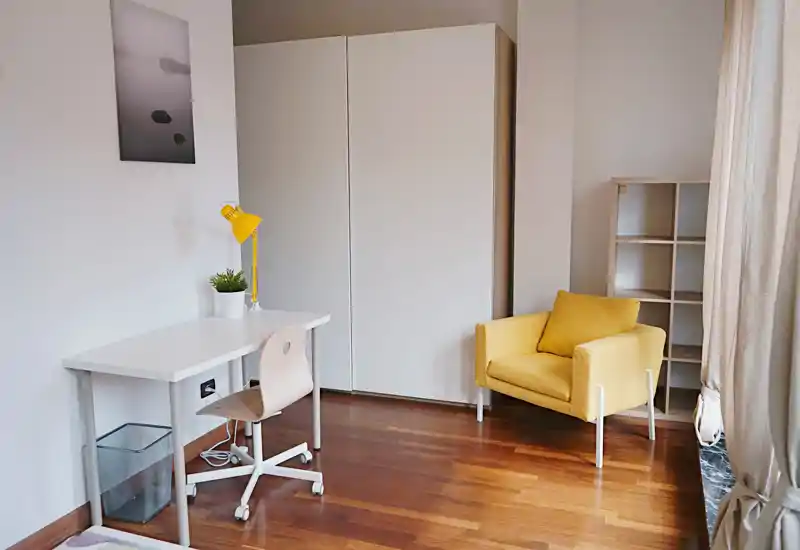 Rincón de dormitorio luminoso y minimalista con armario blanco, escritorio y sillón amarillo; suelo de madera y luz natural que generan una atmósfera serena.