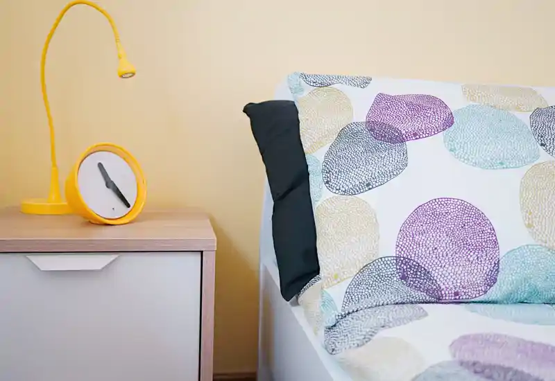 Rincón de dormitorio luminoso y moderno con ropa de cama estampada, almohada oscura y mesita con lámpara y reloj amarillos — ideal para una habitación individual o de invitados.