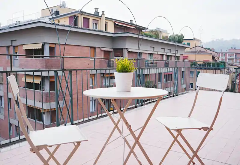 Terraza soleada con una mesa redonda pequeña, dos sillas plegables y una planta en maceta, con vistas a edificios de ladrillo — ideal para el café de la mañana.