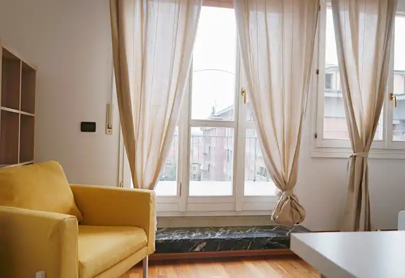 Zona de estar luminosa y acogedora con un sillón amarillo cómodo, cortinas suaves y una gran ventana que aporta luz natural — ideal para relajarse o leer.