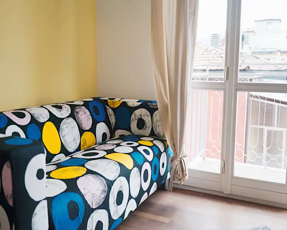 Rincón de salón moderno y luminoso con un sofá colorido de motivos geométricos junto a grandes ventanas que aportan abundante luz natural.