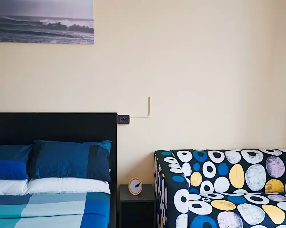 Rincón de dormitorio luminoso con cama bien hecha, cabecero oscuro y ropa de cama azul, sofá colorido con estampado y mesita de noche — perfecto para una estancia acogedora.