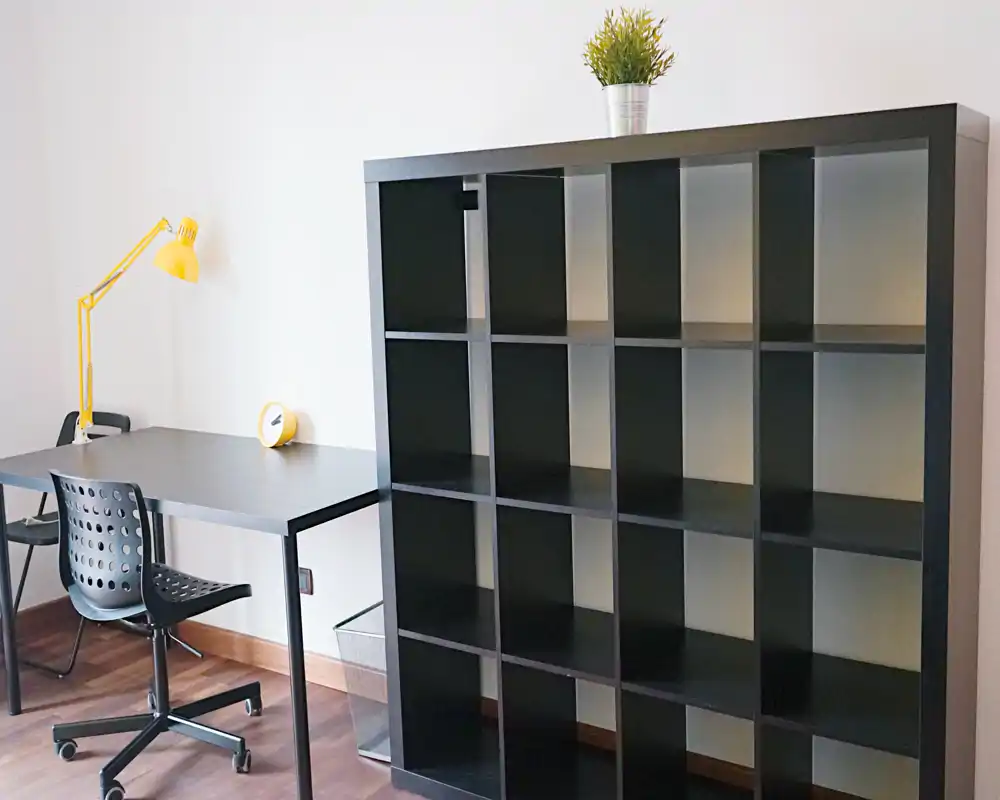 Rincón de trabajo luminoso y minimalista con escritorio negro, silla ergonómica, lámpara amarilla y gran estantería de cubos — ideal para una oficina en casa compacta o zona de estudio.